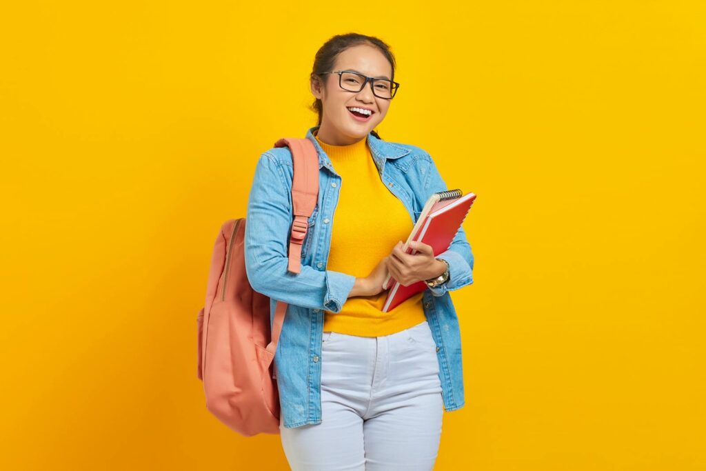 Cheerful student in denim and backpack holding notebooks against a yellow background.